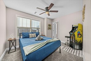 A bedroom with a bed, a ceiling fan, a window with blinds, a door, and a Pac-Man arcade machine. - Month-to-Month Rental (Corporate Stays) (Clarksville)