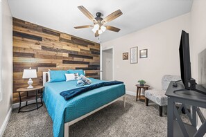 A bedroom with a wooden headboard, a bed with teal bedding, a ceiling fan, a desk with a television, and a small plant. - Month-to-Month Rental (Corporate Stays) (Clarksville)