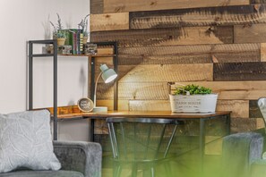 A cozy seating area with a wooden wall, a metal shelf with books and plants, and a table with a lamp and a planter. - Month-to-Month Rental (Corporate Stays) (Clarksville)