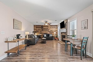 A modern living room with a wooden floor, a ceiling fan, a fireplace, and a dining area with a table and chairs. - Month-to-Month Rental (Corporate Stays) (Clarksville)