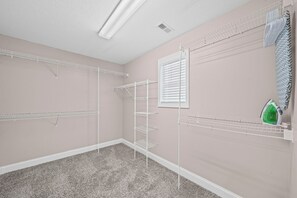 A room with a pink wall, a white ceiling, and a carpeted floor. There are white wire shelves on the walls and a green iron on a shelf. - Month-to-Month Rental (Corporate Stays) (Clarksville)