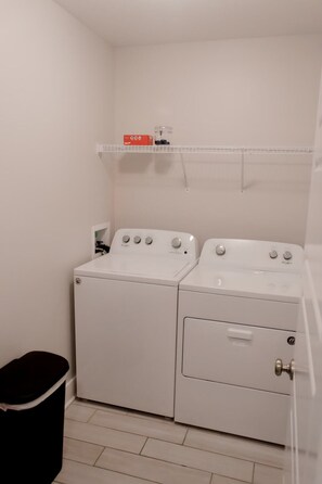 A laundry room with two white Whirlpool washers, a black trash bin, and a shelf with a red box. - The Lookout, by Lori in Downtown (88 Promontory) (Clarksville)
