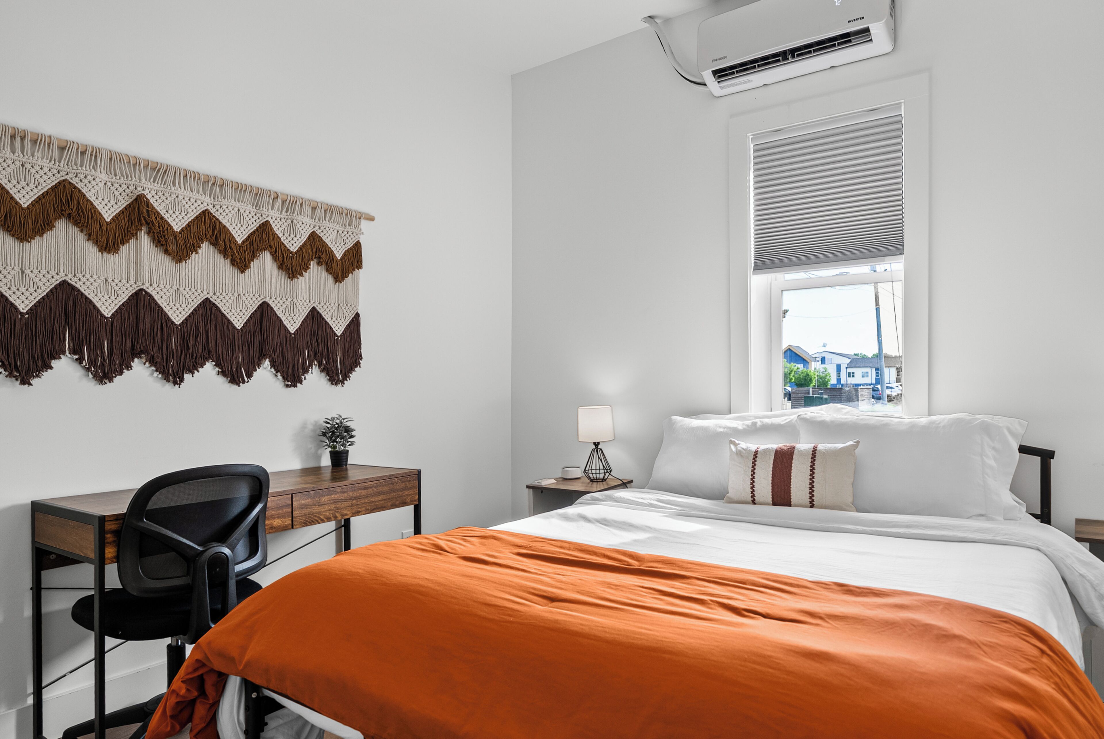 A bedroom with a bed, a desk, a chair, a lamp, and a window with blinds.