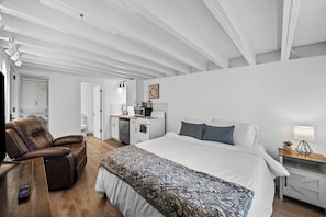A bedroom with a bed, a reclining chair, a desk, a lamp, and a small kitchenette.