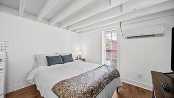 A bedroom with a bed, bedside table, lamp, air conditioning unit, and a window with blinds.
