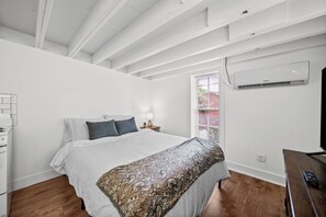 Une chambre à coucher comprenant un lit, une table de chevet, une lampe, un appareil de climatisation et une fenêtre avec des stores.