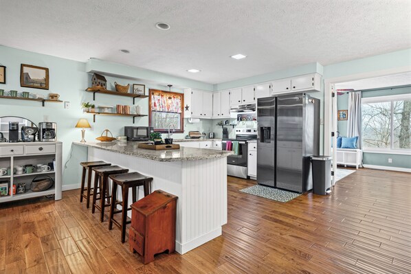 A modern kitchen with white cabinetry, a large island with stools, a stainless steel refrigerator, and a window with a view.