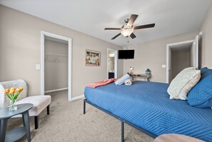 A bedroom with a bed, a ceiling fan, a TV, a chair, and a table with a vase of flowers.