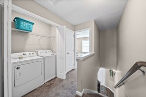 A laundry room with two white washers and dryers, a blue laundry basket on a shelf, and a small window with blinds.