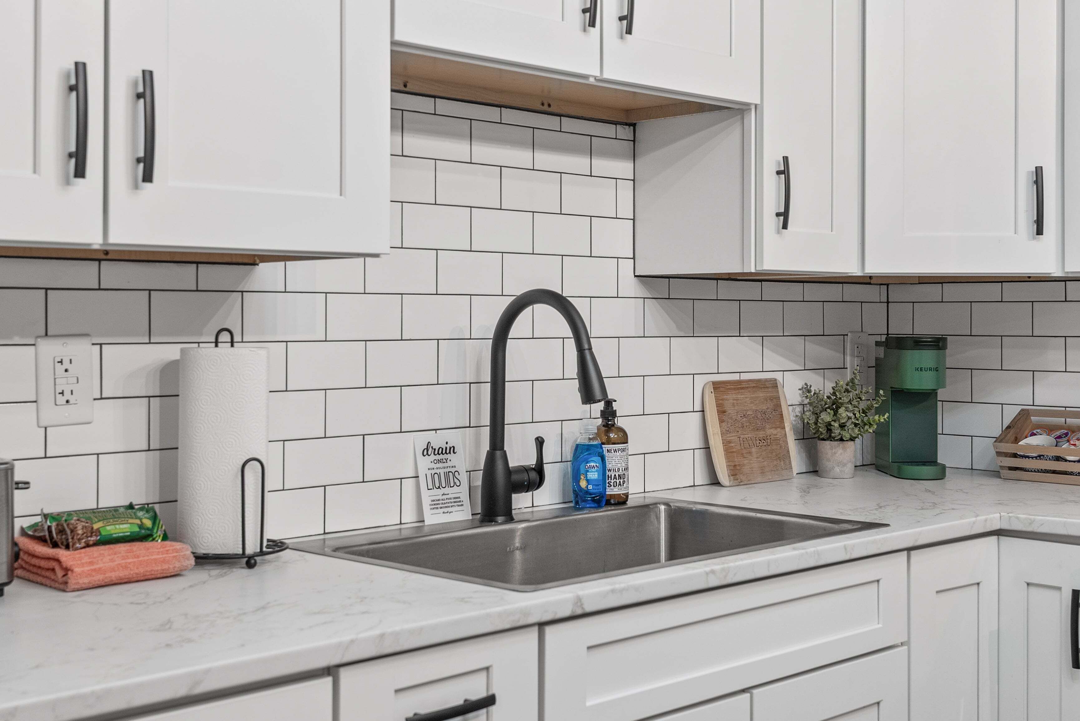 Une cuisine avec des armoires blanches, un évier en acier inoxydable, un porte-serviettes en papier, un distributeur de savon et une machine à café Keurig.