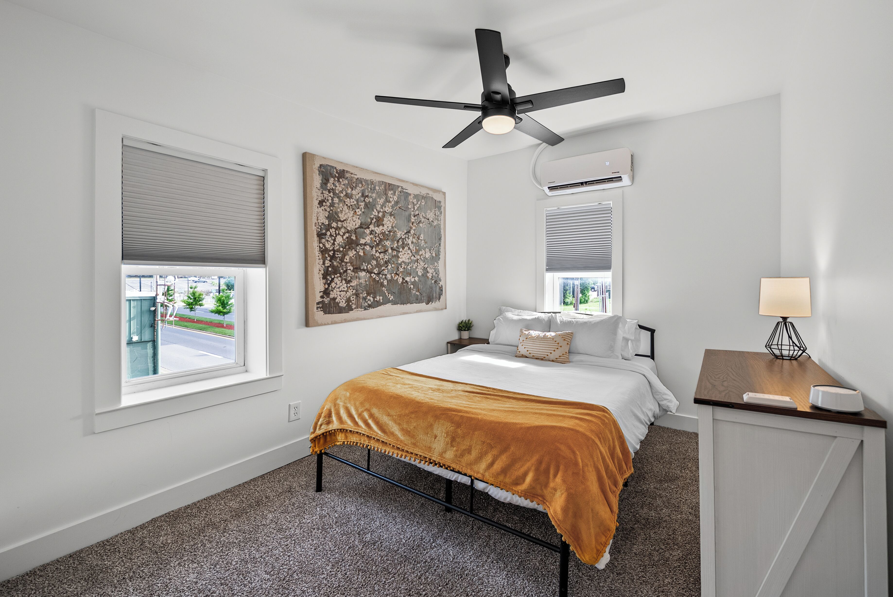 A bedroom with a bed, a ceiling fan, a window with blinds, a painting on the wall, and a bedside table with a lamp.