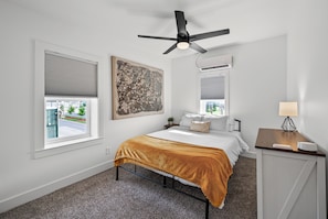 A bedroom with a bed, a ceiling fan, a window with blinds, a painting on the wall, and a bedside table with a lamp.