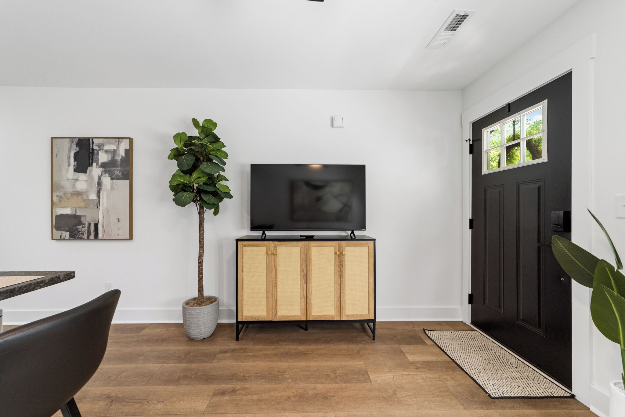 A modern living room with a flat-screen TV on a wooden cabinet, a potted plant, and a dining area with a chair.