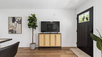 A modern living room with a flat-screen TV on a wooden cabinet, a potted plant, and a dining area with a chair.