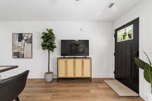 A modern living room with a flat-screen TV on a wooden cabinet, a potted plant, and a dining area with a chair.