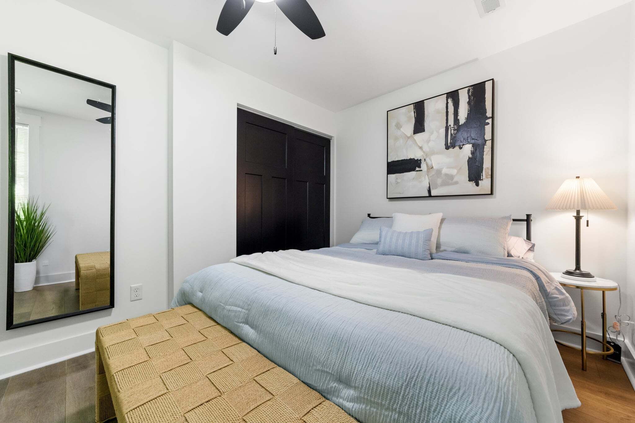 A bedroom with a bed, a bedside table, a lamp, a mirror, a door, and a painting on the wall.