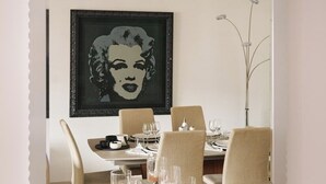 Uma sala de jantar com uma mesa posta para quatro pessoas, uma obra de arte emoldurada acima da mesa e um espelho refletindo o ambiente.