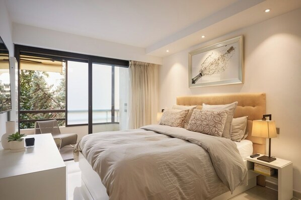 Una camera da letto moderna con un letto grande, una scrivania e una sedia. Si vede una finestra con tende, un quadro appeso al muro e un comodino con una lampada.