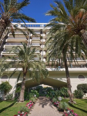Un hotel a più piani con balconi, palme e un giardino curato.