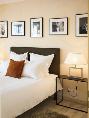 Ein ordentlich bezogenes Bett mit weißen Laken und einem braunen Zierkissen, ein Nachttisch mit Lampe sowie gerahmte Bilder an der Wand.