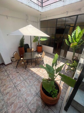 Ein überdachter Patio-Bereich mit Tisch, Stühlen, Topfpflanzen und einem großen Sonnenschirm.