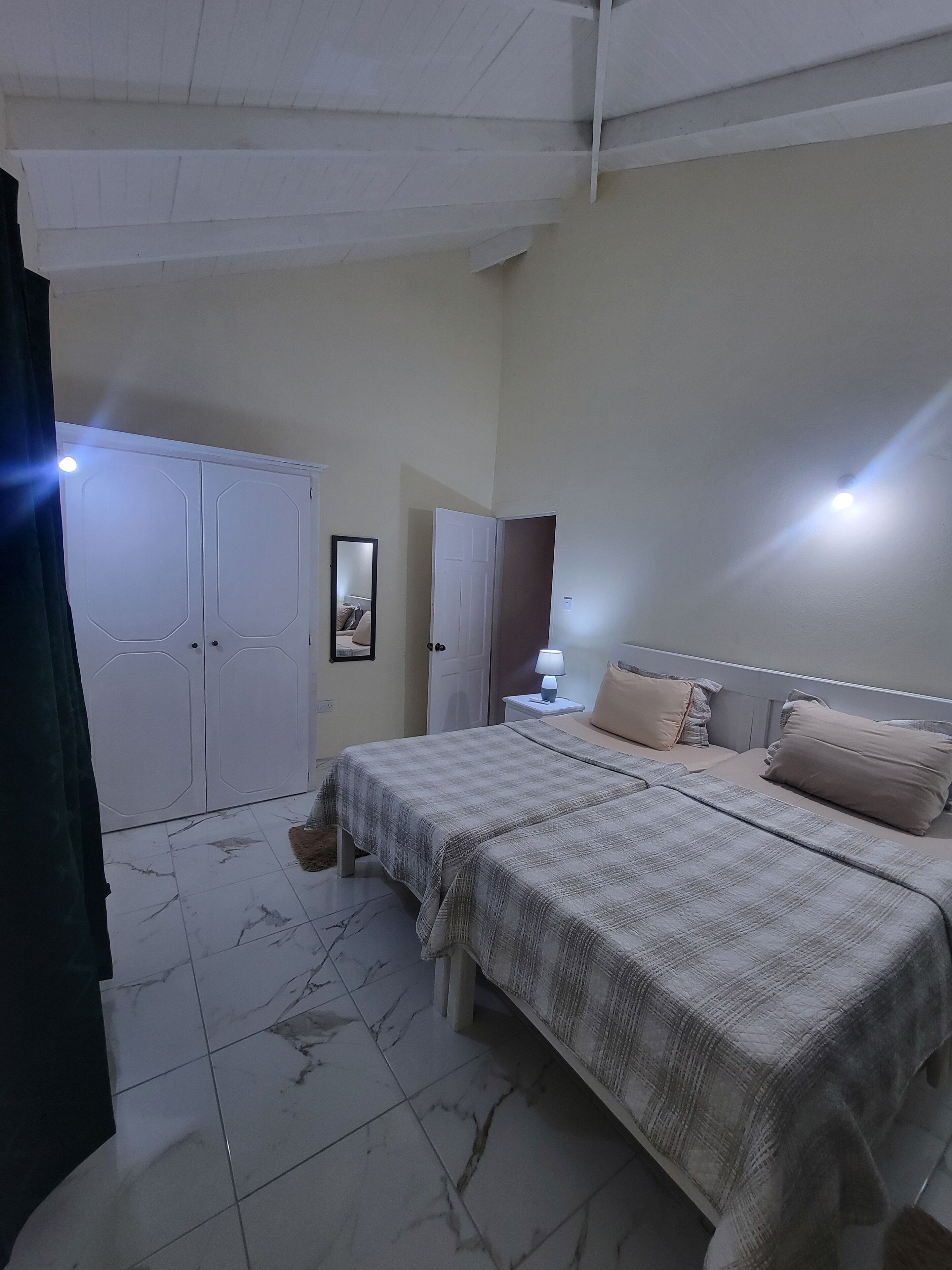 A bedroom with two beds, a wardrobe, a bedside table with a lamp, and a mirror on the wall.