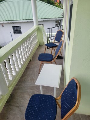 A balcony with two chairs and a small table, a white railing, and a building with a green roof in the background. - ​Country Green — A Peaceful Saint Lucian Countryside Getaway (Castries)