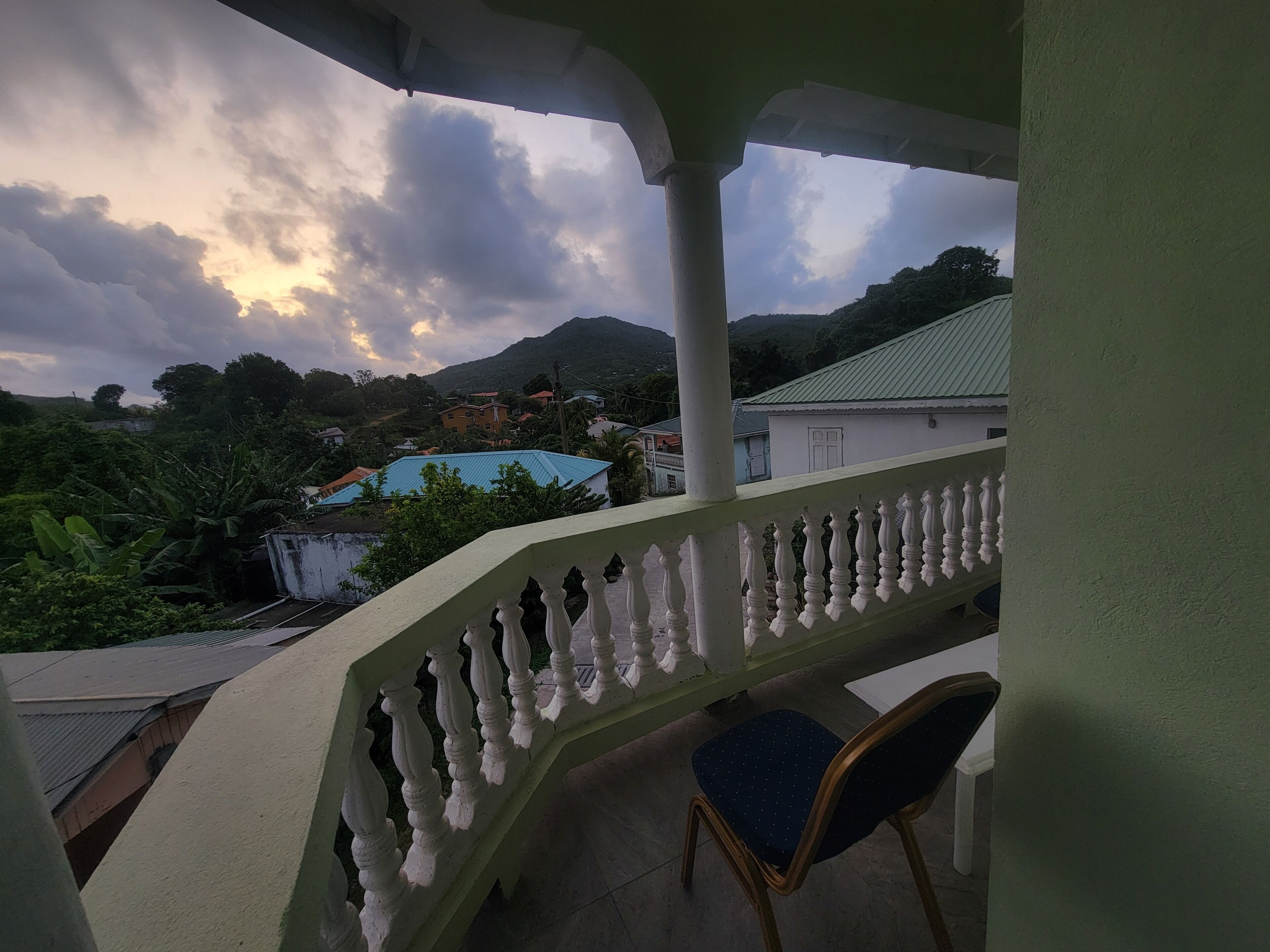 A balcony with a white balustrade, a blue chair, and a view of houses and mountains in the background.