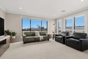 A living room with a sofa, two armchairs, a coffee table, and a TV. There are large windows with blinds, a ceiling light, and a carpeted floor.