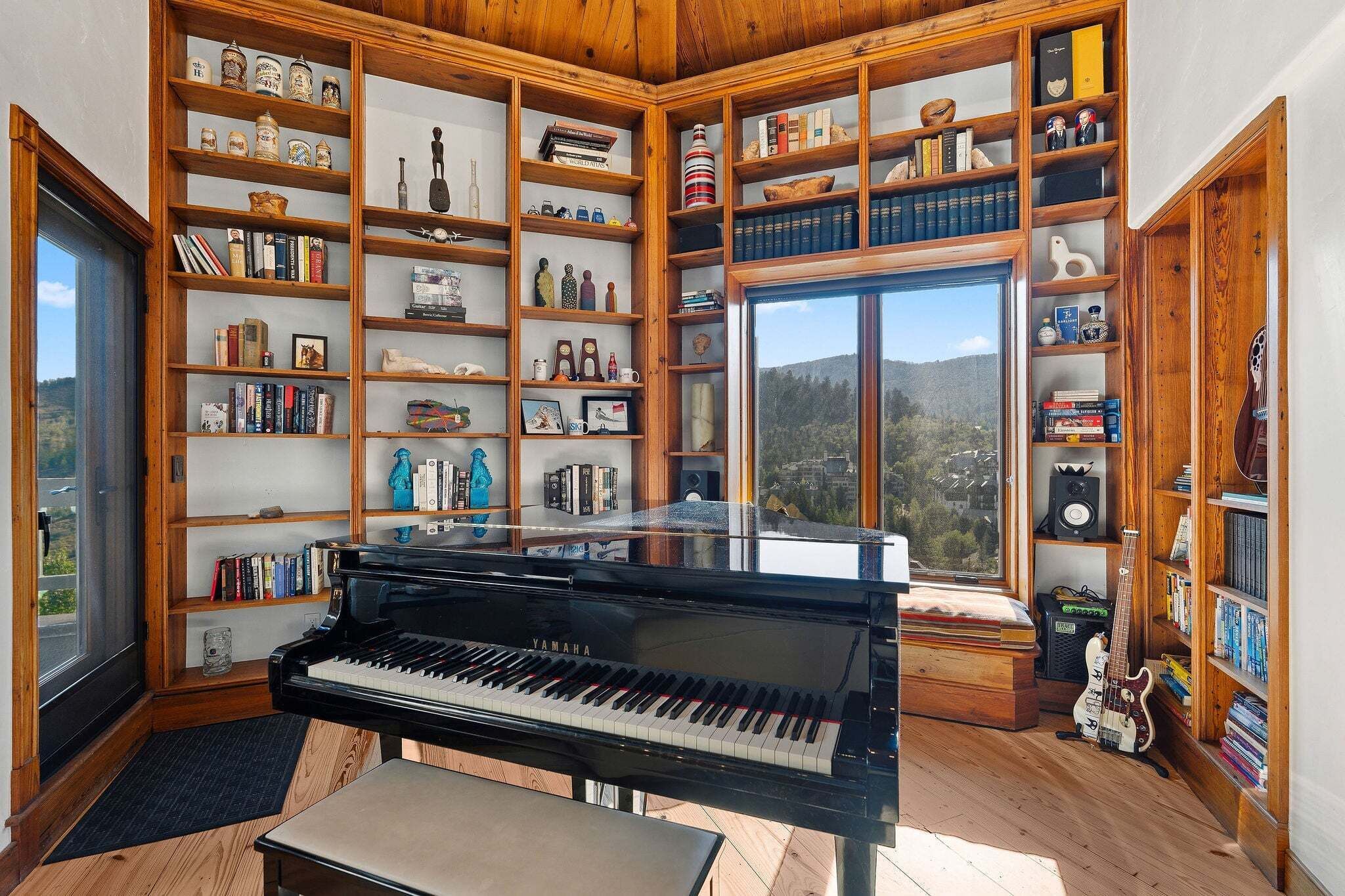 Una habitación con un piano de cola, estanterías de madera llenas de libros y objetos decorativos, y un ventanal con vistas al exterior.