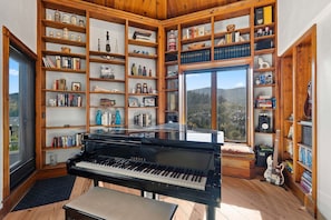 Ein Zimmer mit einem Flügel, Holzregalen voller Bücher und Dekorationsobjekten sowie einem großen Fenster mit Blick ins Freie.