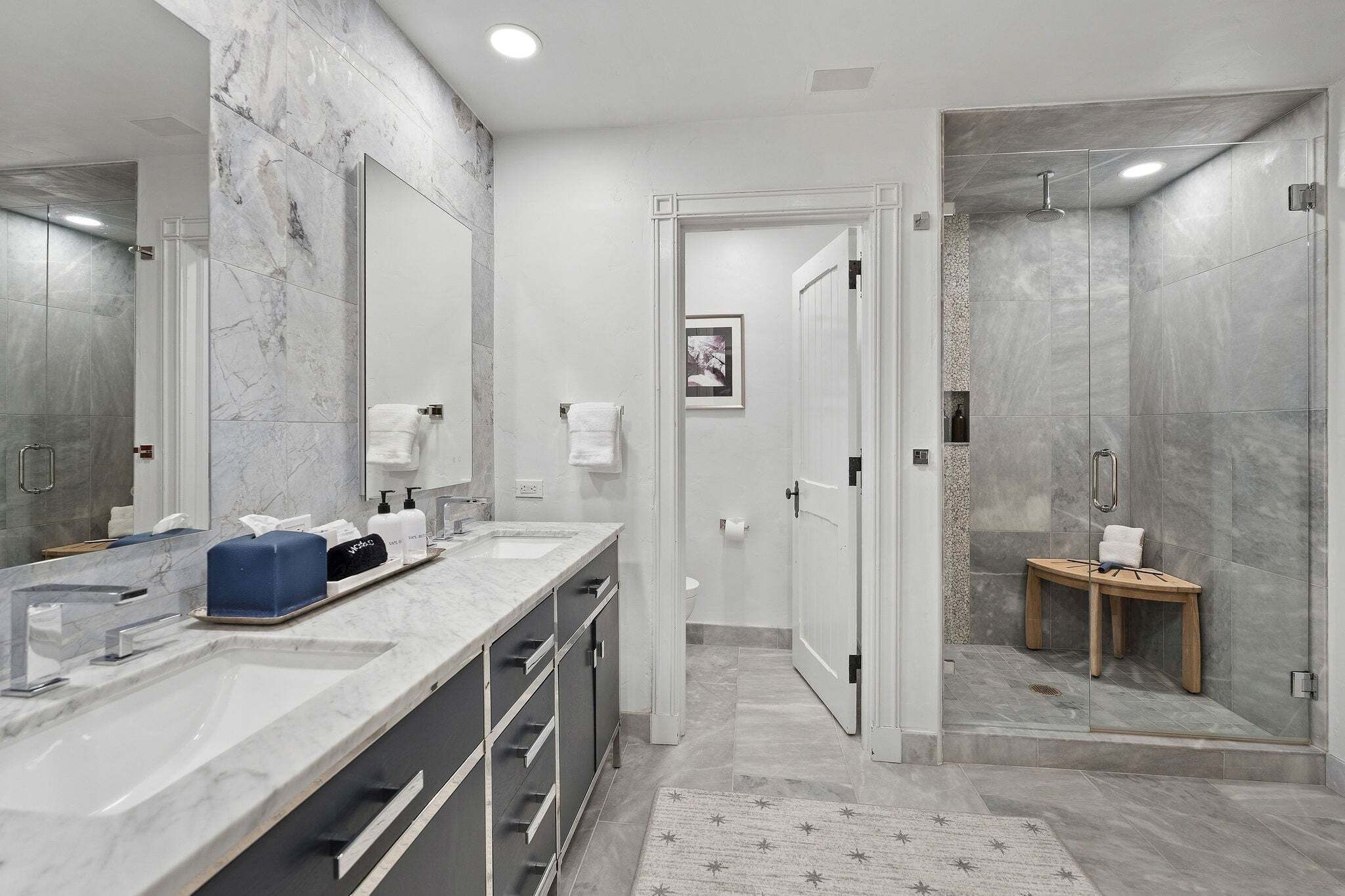 Un baño moderno con tocador de mármol, zona de ducha y un pequeño taburete de madera.