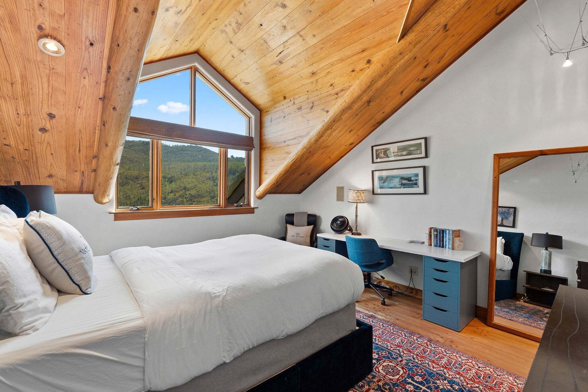 A bedroom with a sloped ceiling, a large window, a bed, a desk, and a chair.