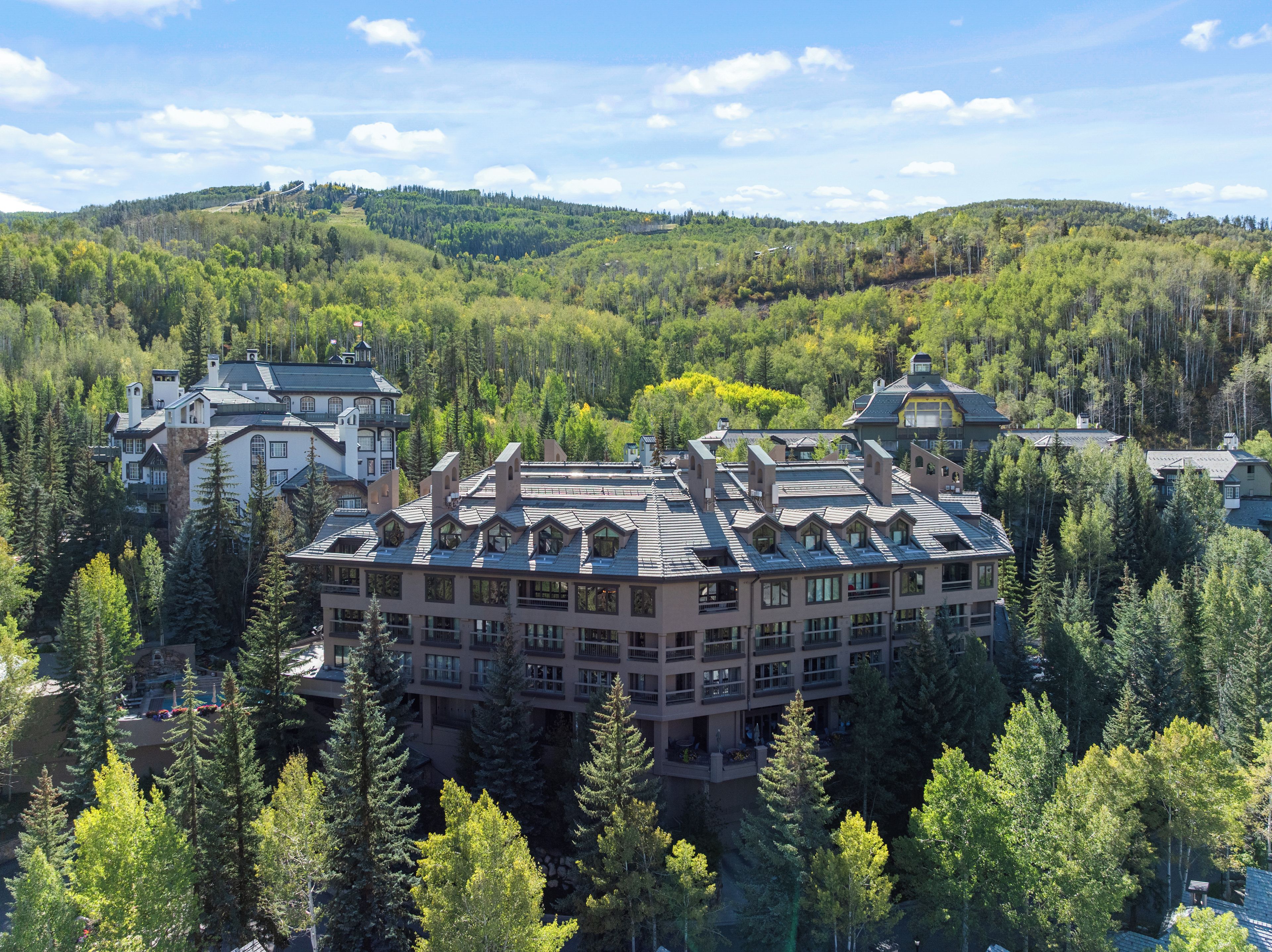 Un grand hôtel à plusieurs étages, entouré d’une forêt dense et avec des collines en arrière-plan.