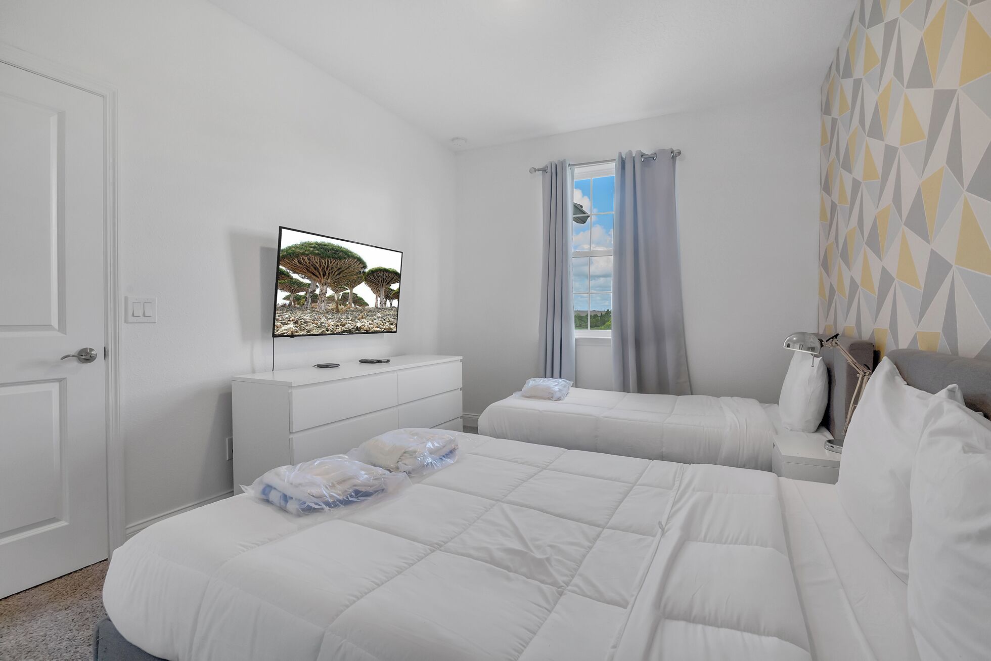 A bedroom with two beds, a TV on the wall, a window with curtains, and a geometric-patterned wall.