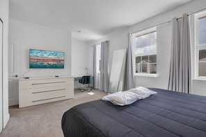 Un dormitorio con cama, escritorio, silla, cómoda, televisor en la pared y ventana con cortinas.