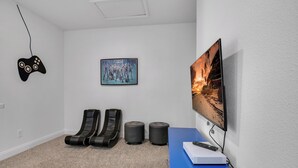 Una habitación con una televisión de pantalla plana grande, un control de videojuegos, dos sillones de masaje, un cuadro y un escritorio azul con un control remoto.