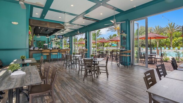 Un comedor interior con mesas y sillas de madera, una zona de bar y grandes ventanales con vistas a una piscina exterior y palmeras.