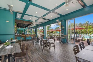 An indoor dining area with wooden tables and chairs, a bar area, and large windows offering views of an outdoor pool and palm trees. - 4BR Family Townhome, pool, resort & Splash (Davenport)