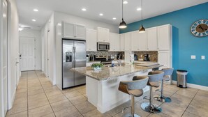 Cocina moderna con armarios blancos, refrigerador de acero inoxidable, microondas integrado y encimera de granito. Hay taburetes de bar y un reloj en la pared.
