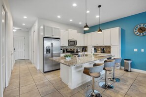 A modern kitchen with white cabinets, a stainless steel refrigerator, a built-in microwave, and a granite countertop. There are bar stools and a clock on the wall. - 4BR Family Townhome, pool, resort & Splash (Davenport)
