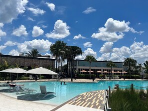 A swimming pool with lounge chairs and umbrellas, surrounded by palm trees and a building with a brick facade. - 4BR Family Townhome, pool, resort & Splash (Davenport)