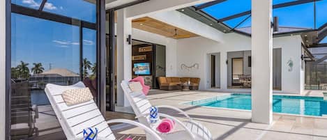 A modern outdoor pool area with two lounge chairs, a television, and a dining area.