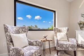 Ein Zimmer mit zwei gemusterten Sesseln, einem kleinen Holztisch und Blick auf einen klaren, blauen Himmel durch ein großes Fenster.