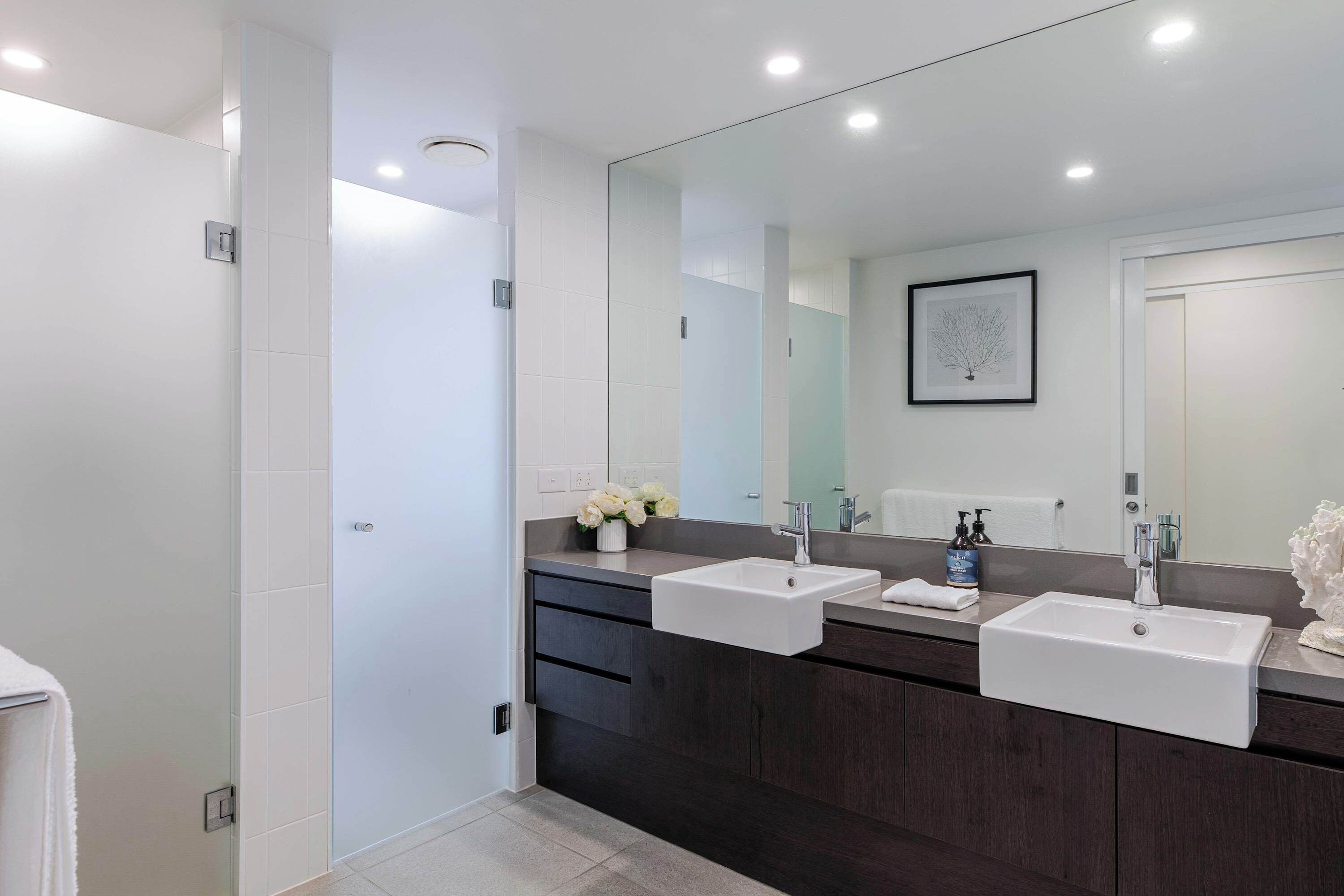 A modern bathroom with two sinks, dark wood cabinets, a large mirror, and a framed artwork on the wall.