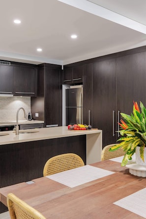 A modern kitchen with dark cabinetry, a stainless steel refrigerator, and a dining area with a wooden table and chairs. - SeaEden @ The Temple 3 Bedroom with Private Pool (Craiglie)