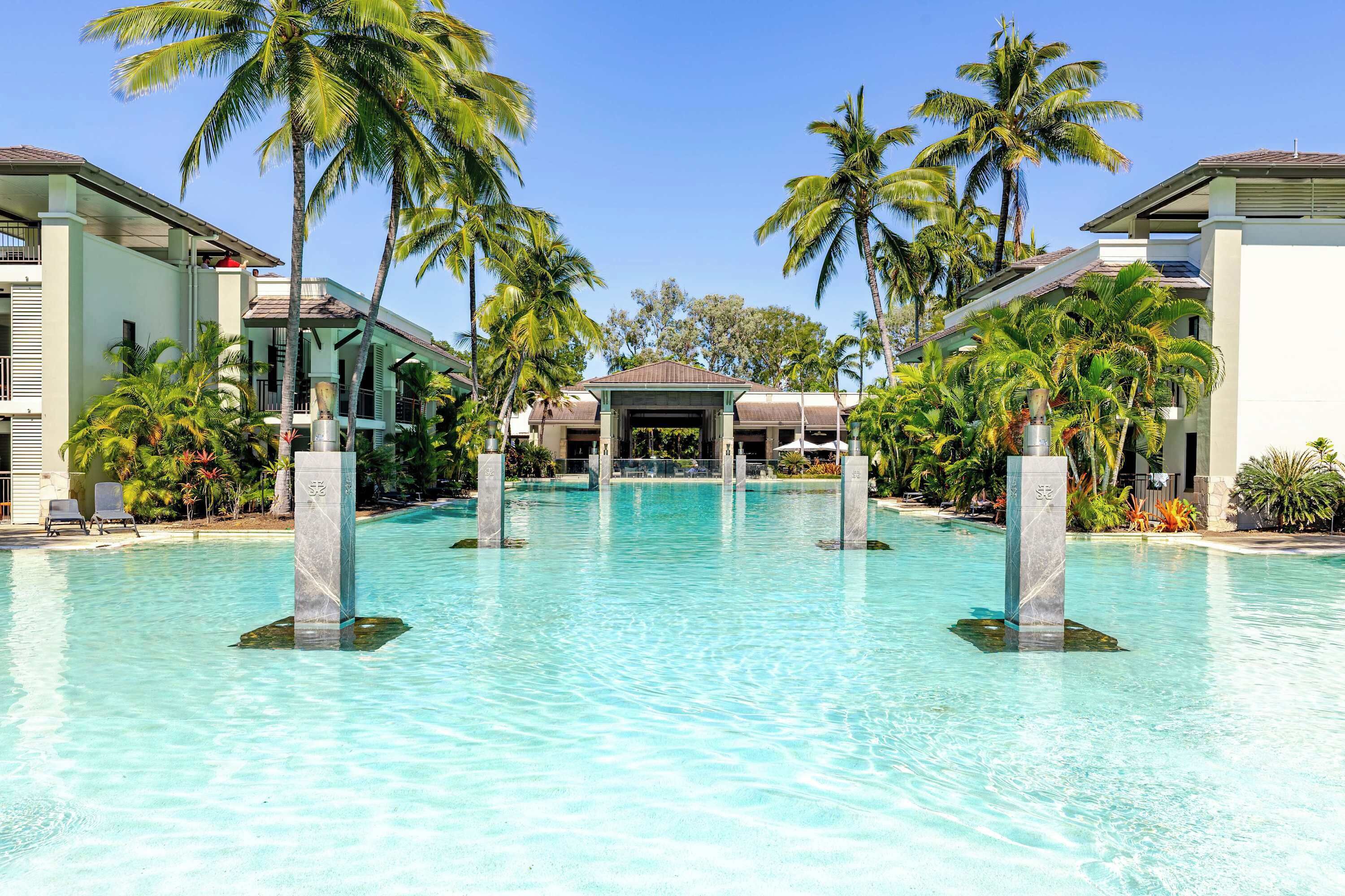 A resort pool with clear water, surrounded by palm trees and modern buildings.