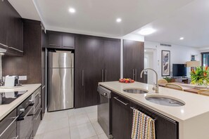 A modern kitchen with dark cabinetry, stainless steel appliances, a central island with a sink, and a dining area with a table and chairs. - SeaEden @ The Temple 2 Bedroom with Private Pool (Craiglie)