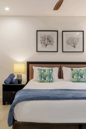 Cama bem arrumada com um cobertor azul, duas almofadas com estampas de folhas tropicais, um criado-mudo com abajur e duas obras de arte com tema de árvores emolduradas na parede.