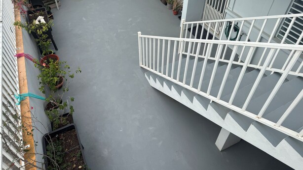 A rooftop terrace with potted plants, a white railing, and a grey floor.